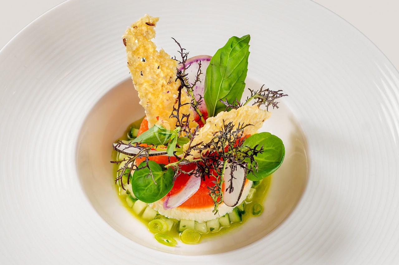 Assiette gastronomique composée de légumes frais, herbes, radis, saumon et tuiles croustillantes sur fond blanc.