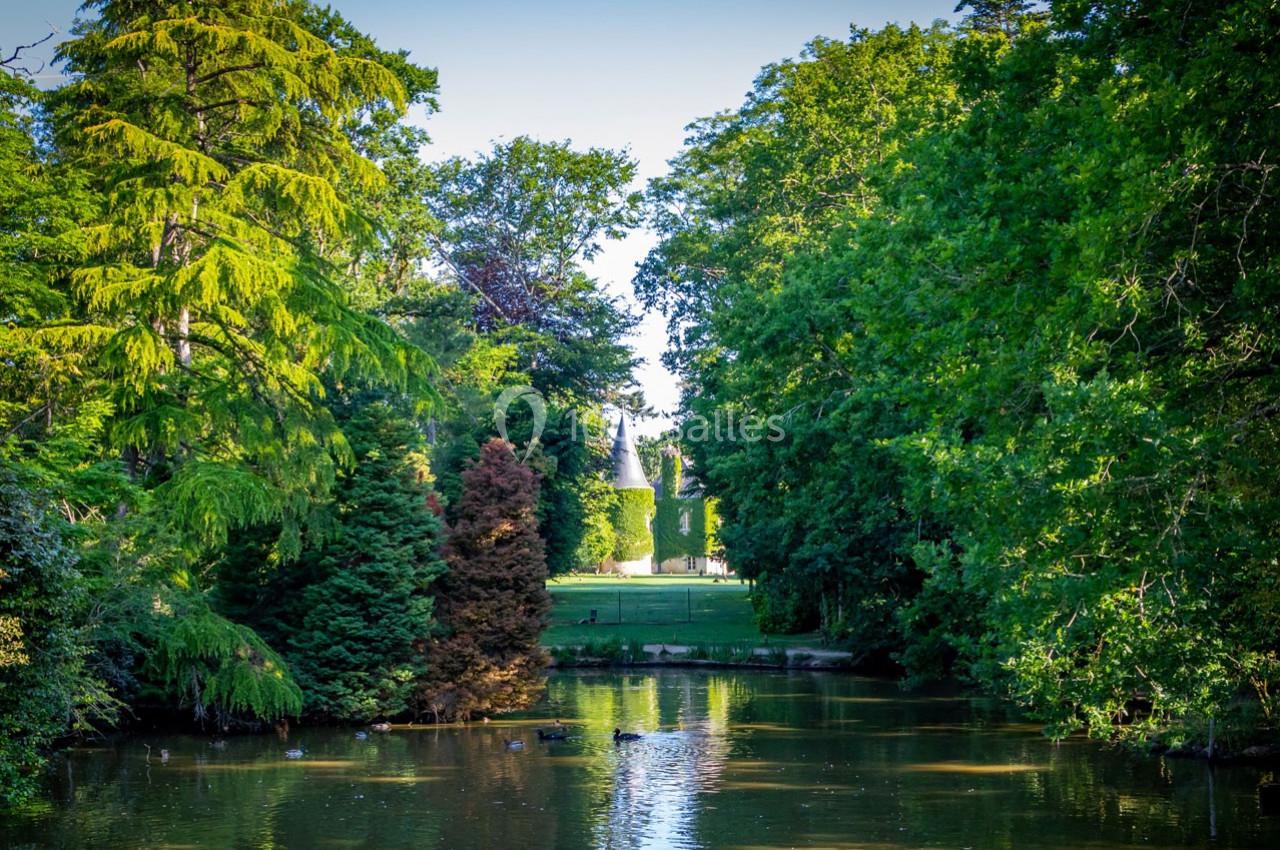 Vue d'un étang entouré d'arbres verdoyants avec une pelouse et un bâtiment visible en arrière-plan.