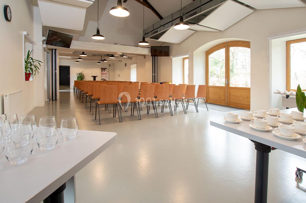 Salle lumineuse avec chaises alignées, tables dressées avec verres et tasses, grandes fenêtres et poutres apparentes.