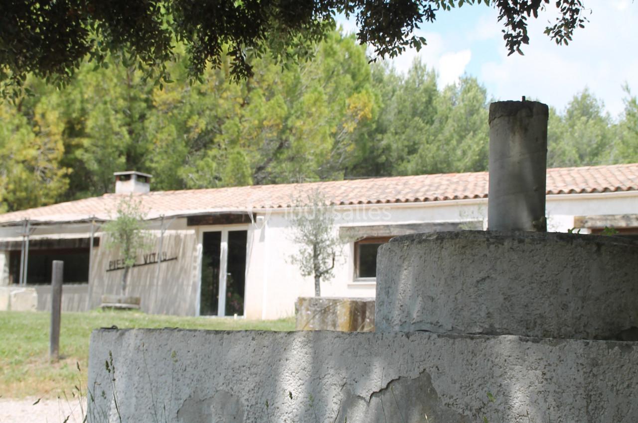 Vue d'un bâtiment rural entouré de végétation, avec un muret en béton au premier plan sous des arbres.