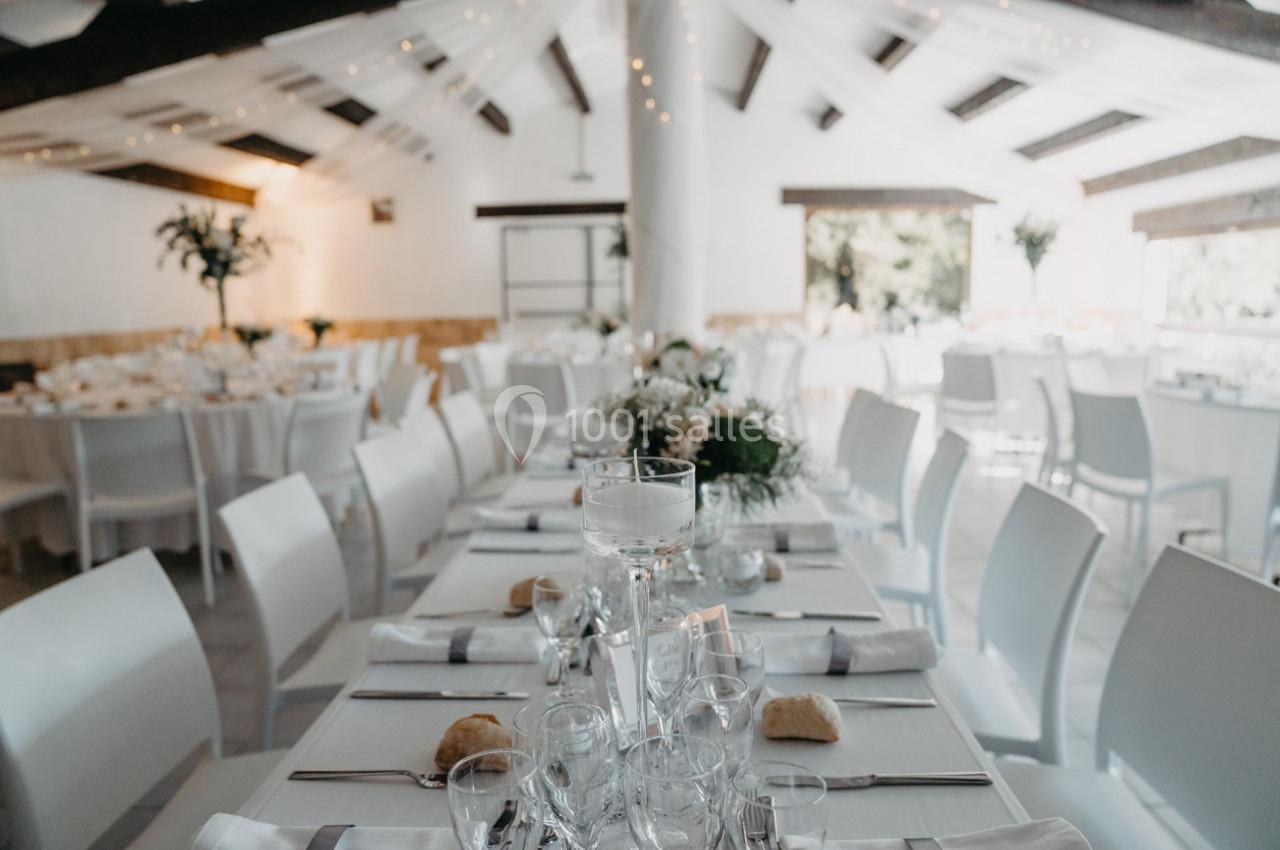Salle de réception lumineuse avec tables dressées, nappes blanches, décorations florales et chaises blanches.