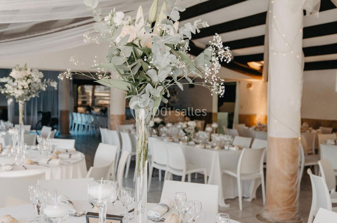 Salle de réception décorée avec des tables rondes, nappes blanches, centres de table floraux et éclairage doux.
