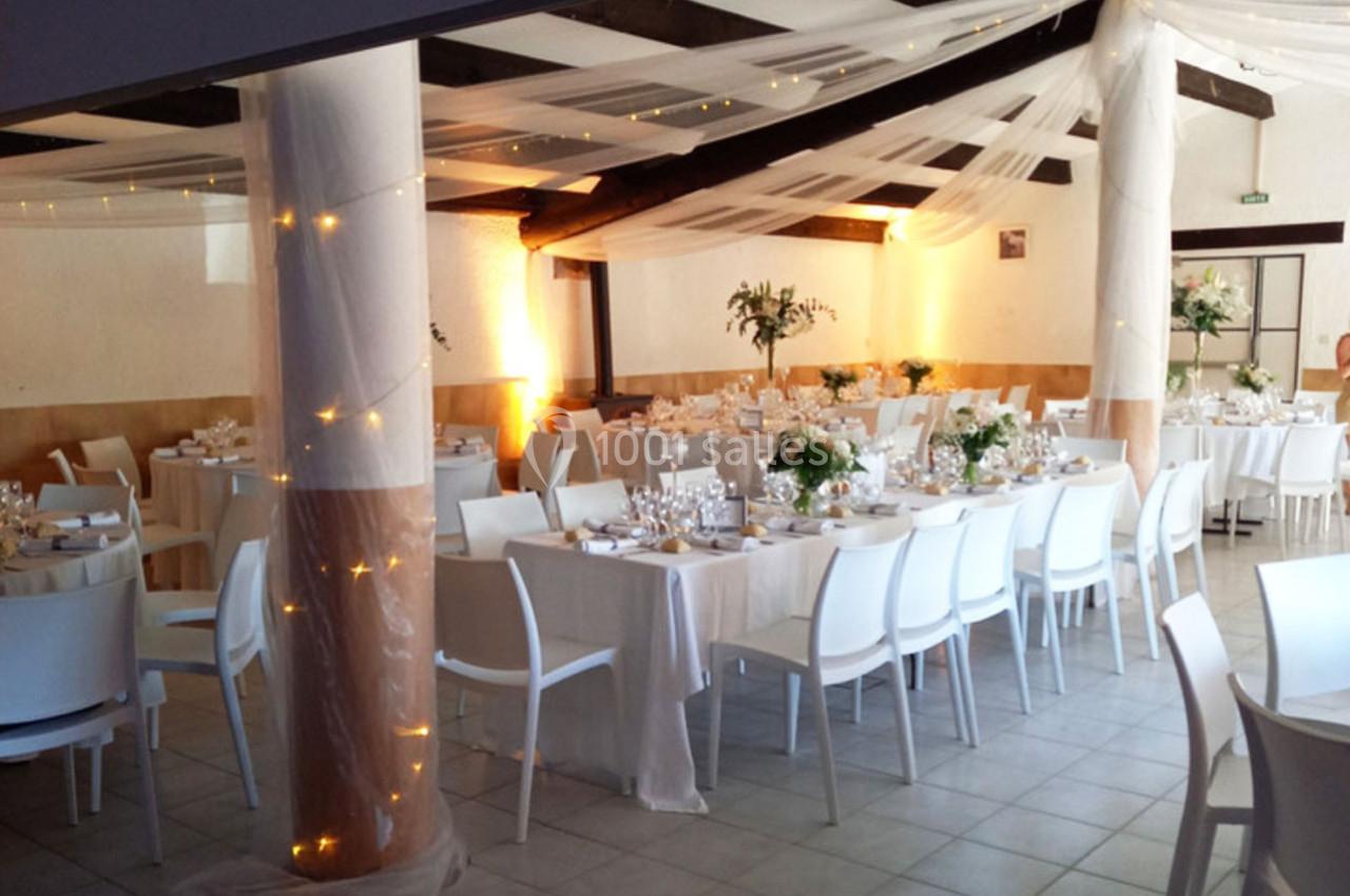 Salle de réception décorée avec des tables dressées, des chaises blanches, des guirlandes lumineuses et des voilages…