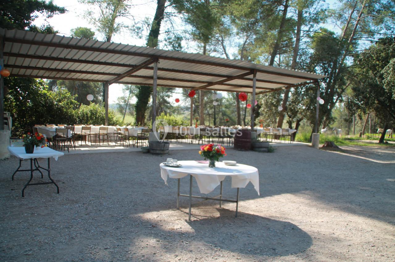 Table dressée en extérieur sous un abri, entourée de chaises, avec des décorations et des arbres en arrière-plan.