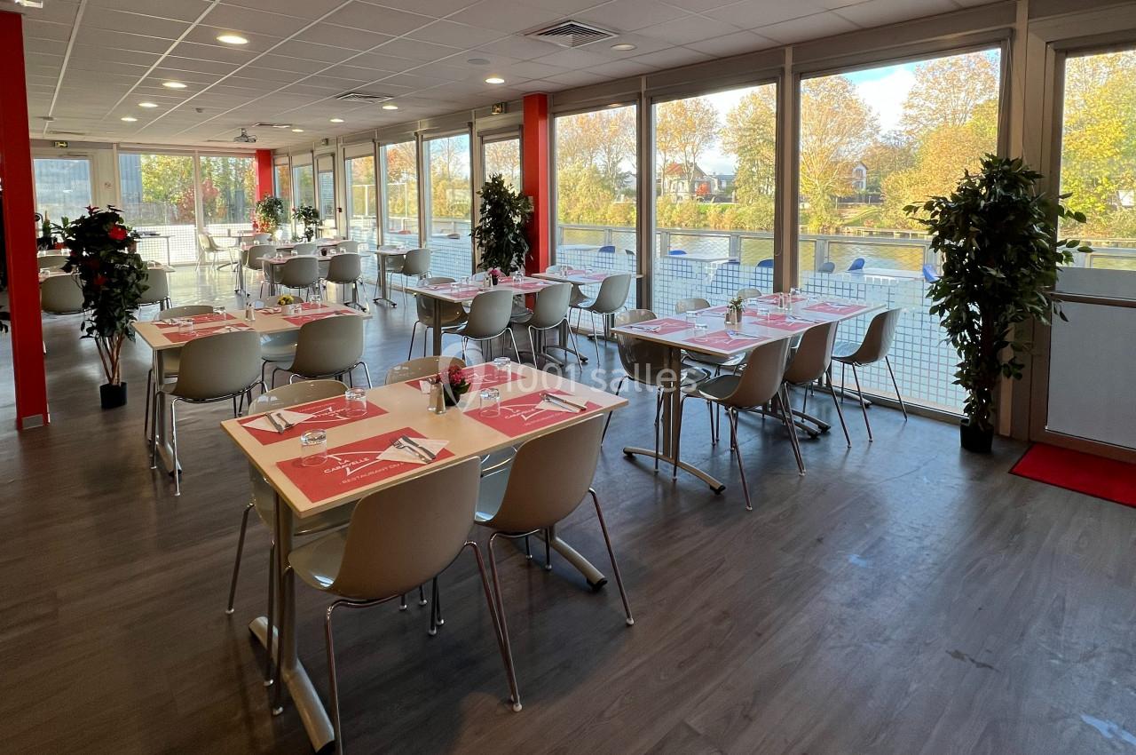 Salle lumineuse avec tables dressées, chaises modernes et grandes baies vitrées donnant sur un paysage arboré.