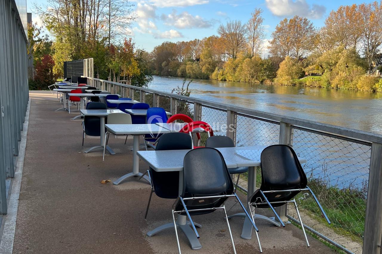 Terrasse avec tables et chaises colorées alignées le long d'une rivière, entourée d'arbres en automne.
