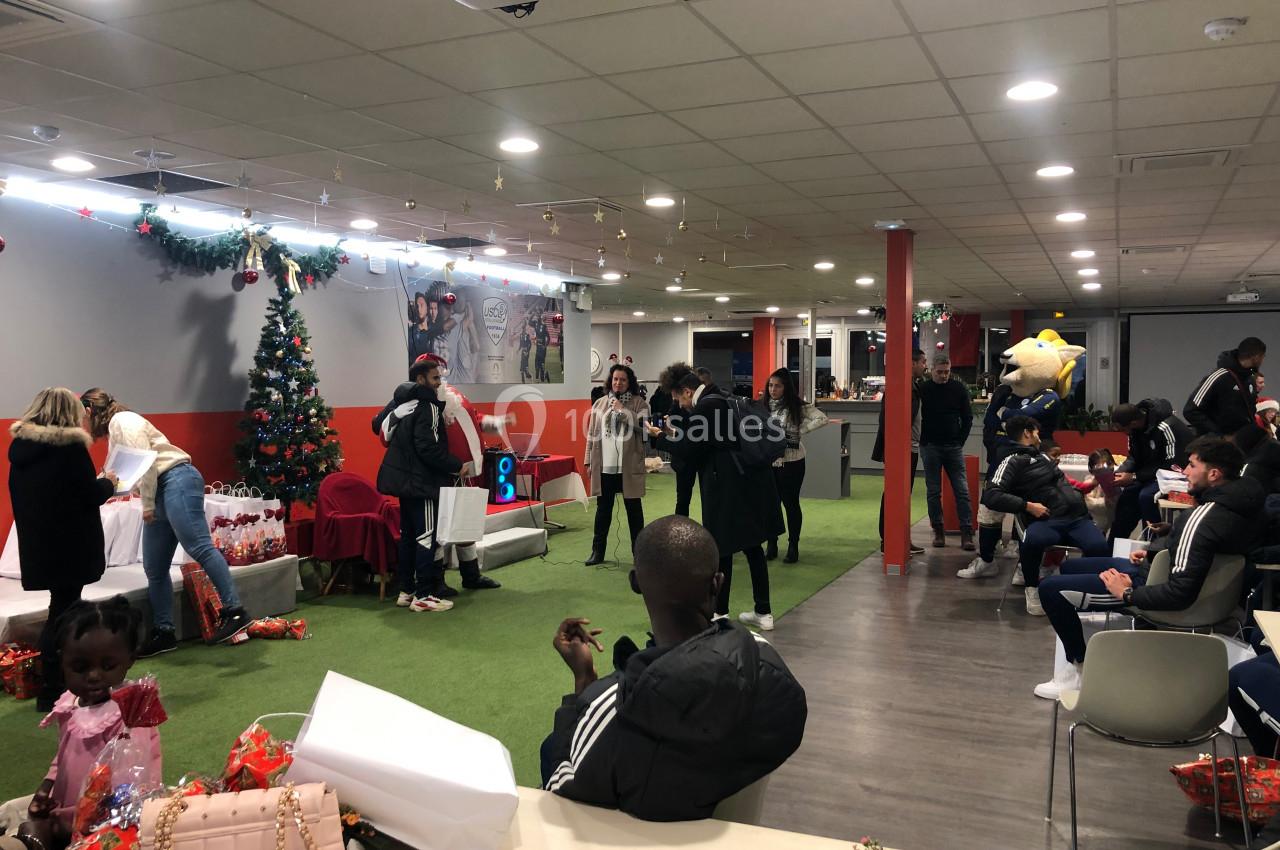 Des personnes participent à une fête dans une salle décorée avec un sapin de Noël et des cadeaux.