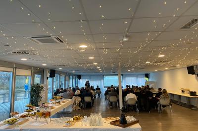 Salle de conférence lumineuse avec des participants assis, une présentation projetée et une table de buffet au premier plan.