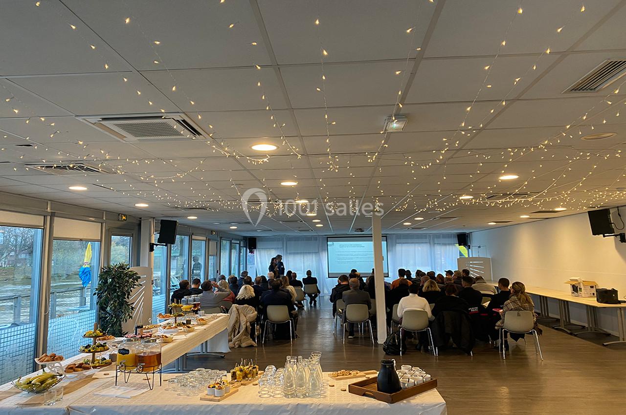 Salle de conférence lumineuse avec des participants assis, une présentation projetée et une table de buffet au premier plan.