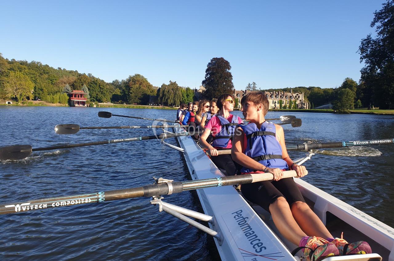 Un groupe de personnes en train de ramer sur un lac calme, entouré de verdure et de bâtiments en arrière-plan.