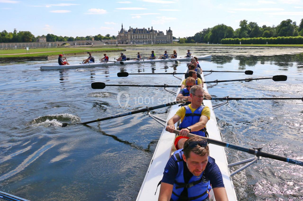 Des rameurs en équipe pagayant sur un plan d'eau calme, avec un château et des arbres en arrière-plan.