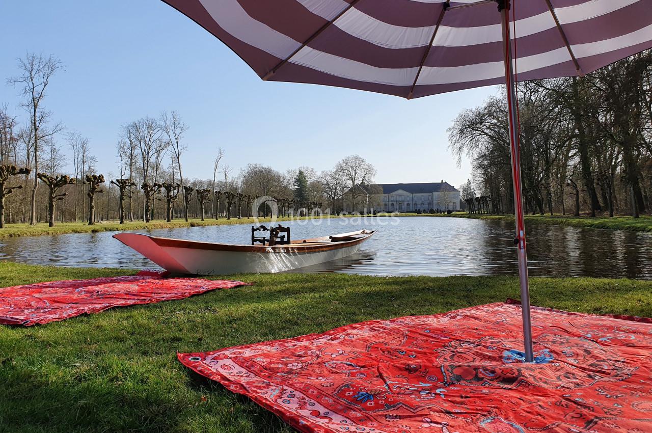 Barque sur un étang entouré d'arbres, avec des tapis rouges et un parasol au premier plan.