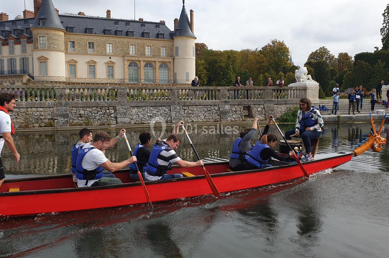 Des personnes pagaient dans une grande barque rouge sur un plan d'eau, près d'un château entouré de verdure.