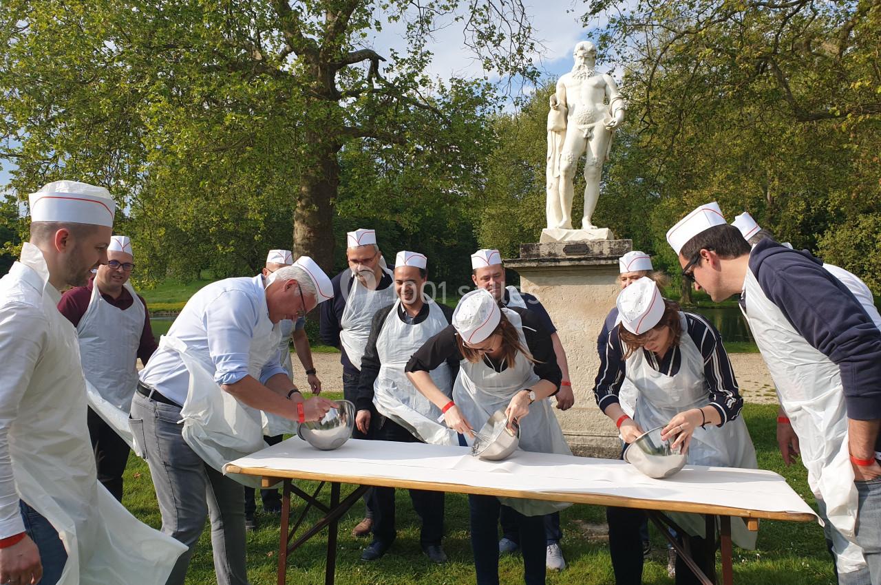 Des personnes en tabliers et toques participent à une activité culinaire en extérieur devant une statue dans un parc.