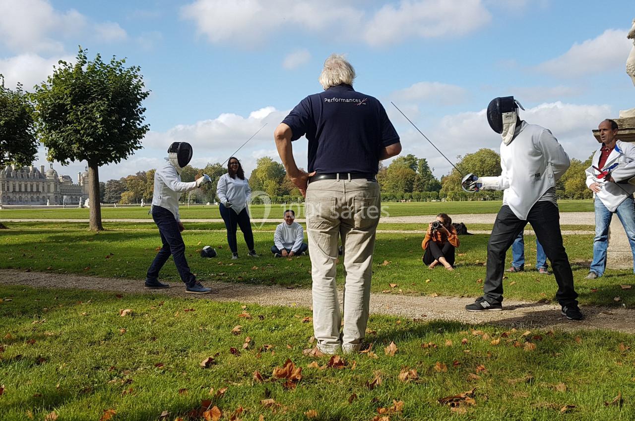 Deux personnes en tenue d'escrime s'affrontent en plein air sous la supervision d'un homme, avec des spectateurs autour.