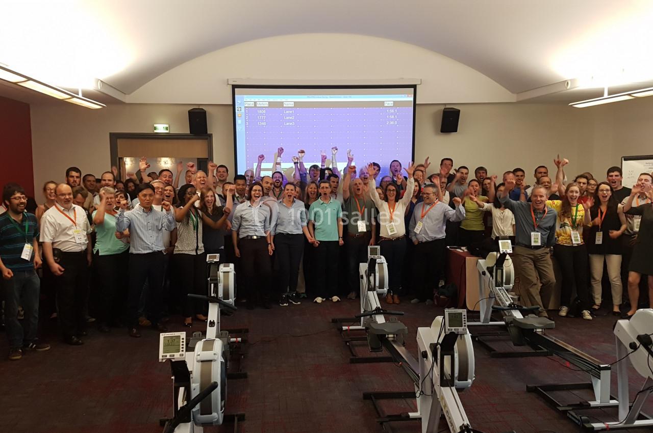 Un groupe de personnes souriantes pose dans une salle avec des rameurs et un écran projeté en arrière-plan.