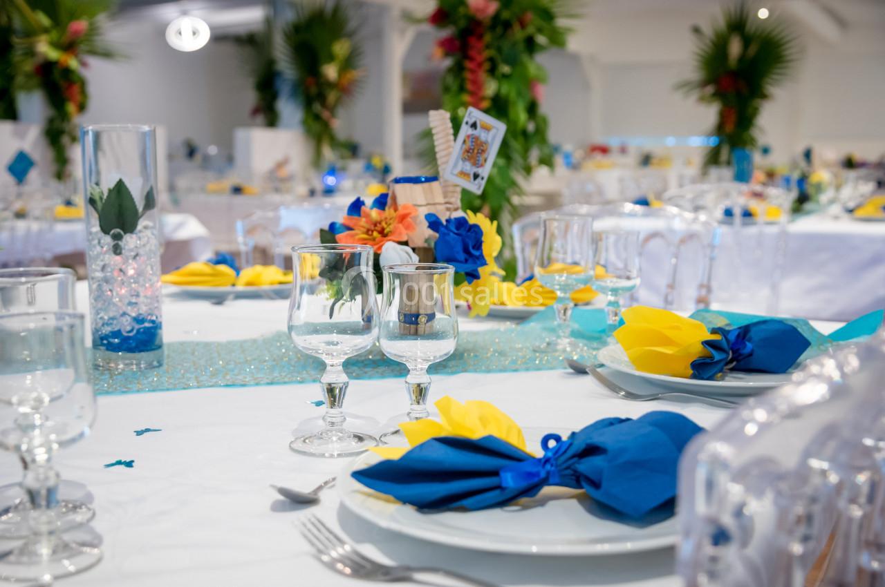 Table dressée avec nappes blanches, serviettes bleues et jaunes, verres et décorations florales colorées.