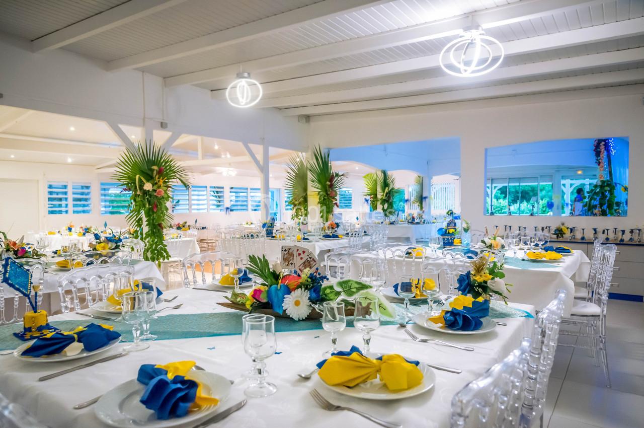 Salle de réception décorée avec des tables dressées, nappes blanches, serviettes jaunes et bleues, et arrangements floraux.