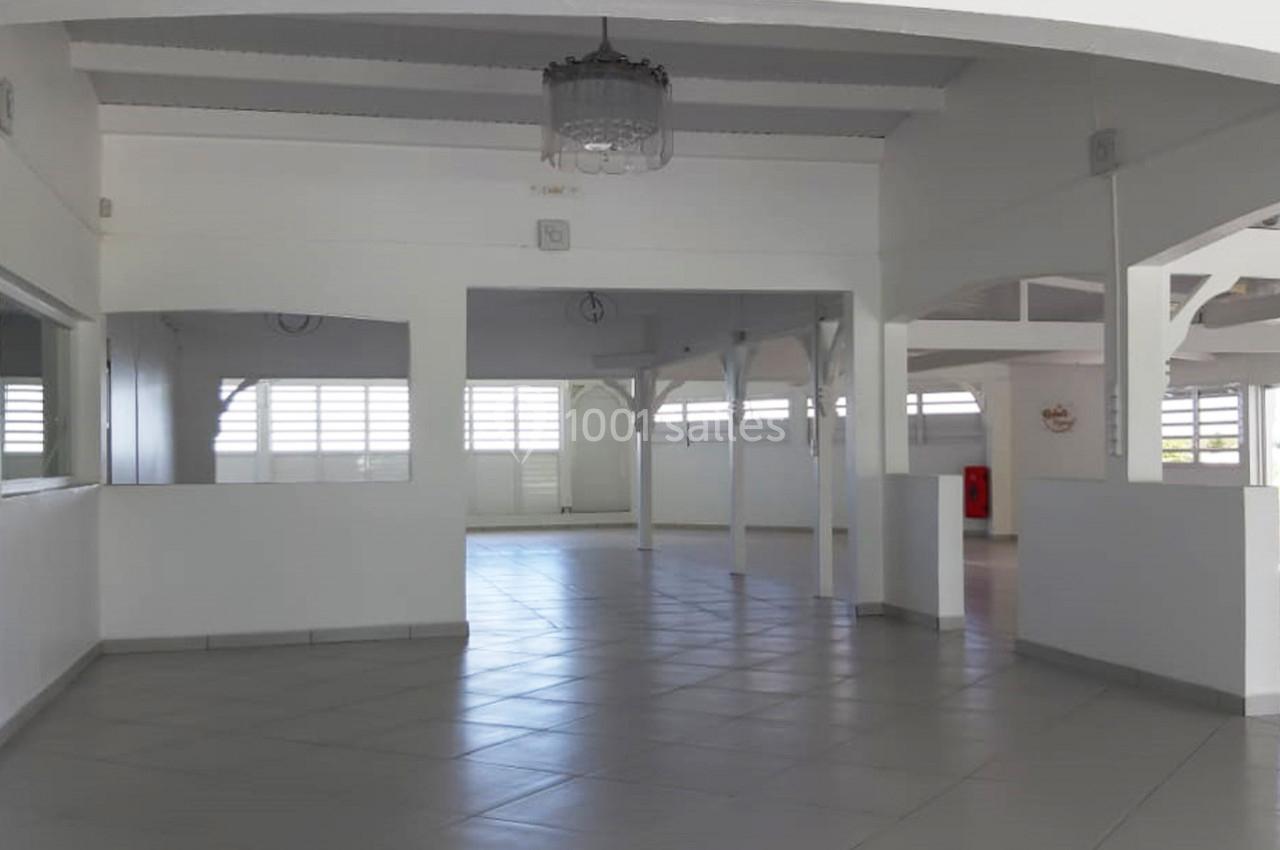 Salle vide lumineuse avec carrelage clair, murs blancs, poutres apparentes et grandes fenêtres laissant entrer la lumière.