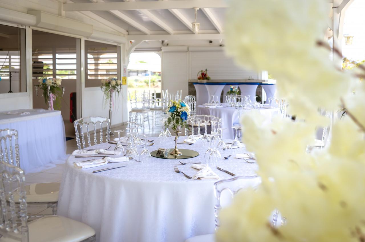 Salle de réception lumineuse avec tables rondes décorées de nappes blanches, fleurs et vaisselle élégante.