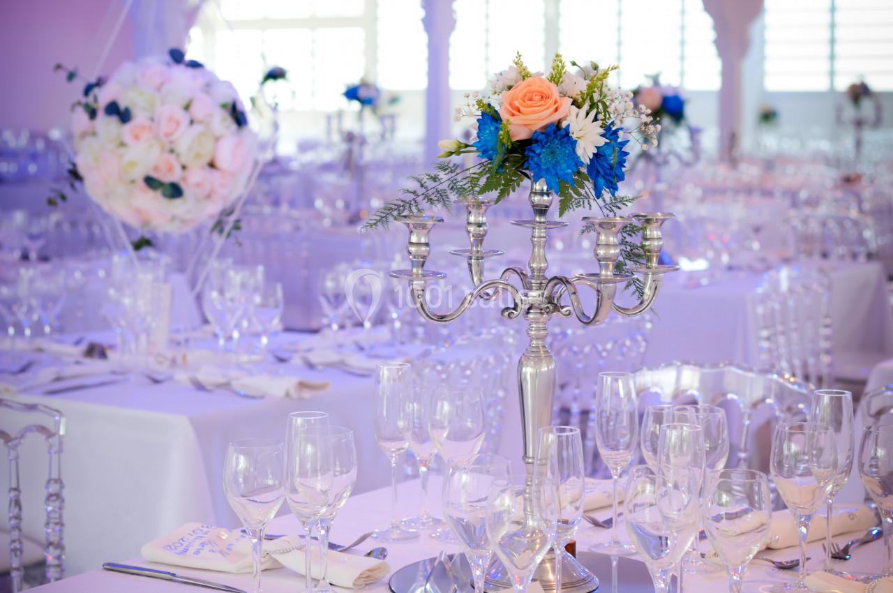 Salle de réception décorée avec des tables dressées, chandeliers argentés et compositions florales aux tons roses et bleus.