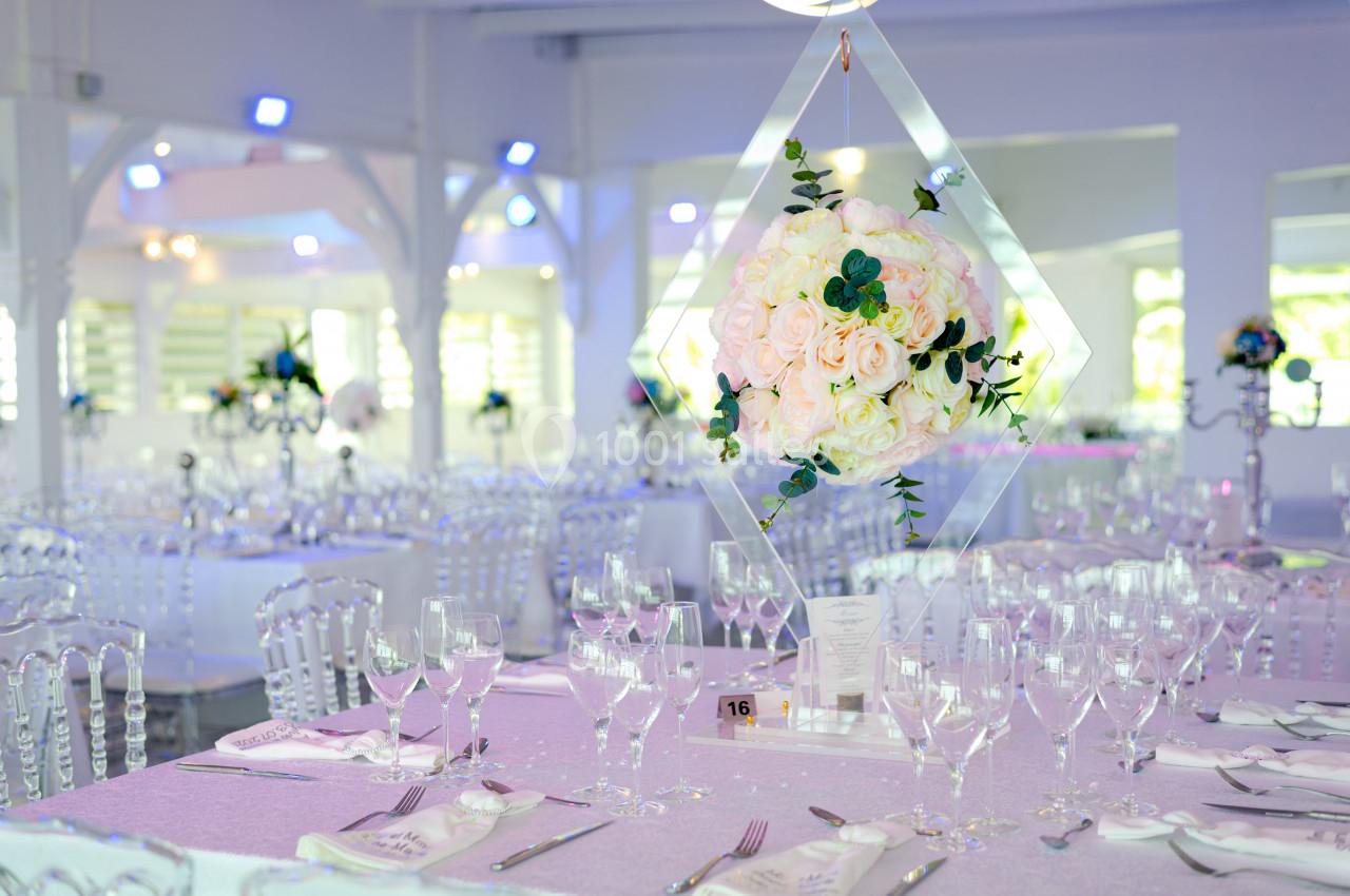 Salle de réception décorée avec des tables élégantes, nappes lilas, chaises transparentes et centre de table floral suspendu.