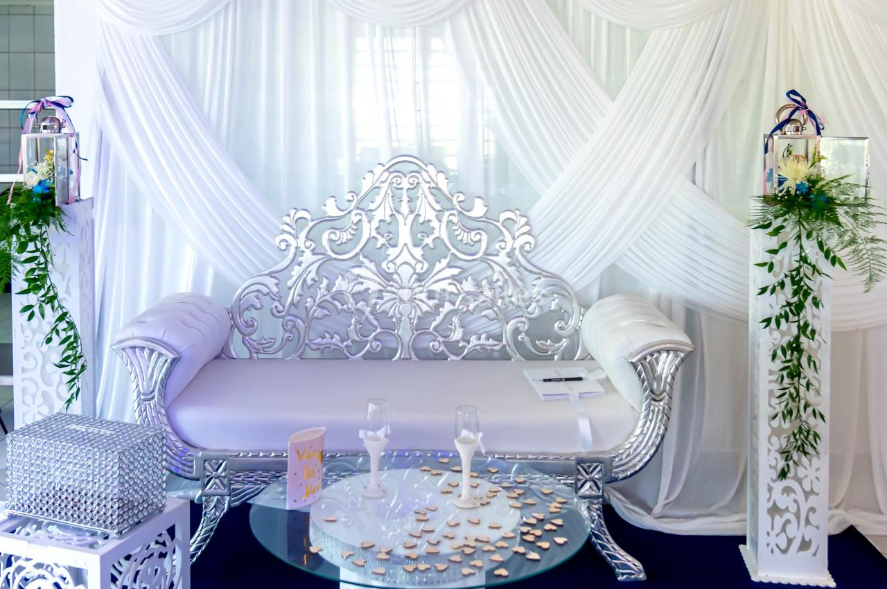 Canapé argenté décoratif devant un rideau blanc drapé, entouré de colonnes fleuries et d'une table basse en verre.