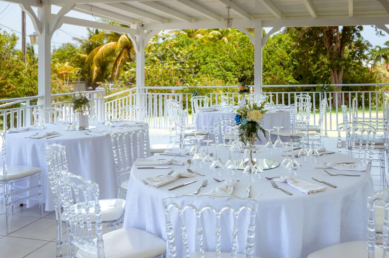 Tables rondes dressées avec nappes blanches et chaises transparentes sous une pergola, entourée de verdure.