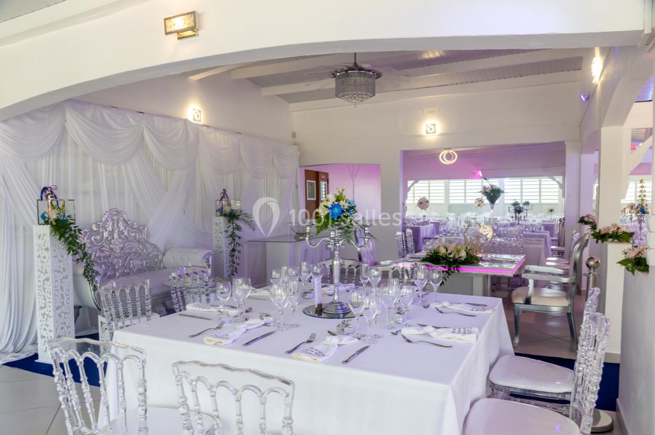 Salle de réception élégante avec tables dressées, nappes blanches, chaises transparentes et décorations florales.