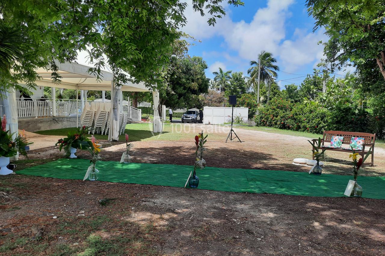 Allée verte décorée de fleurs tropicales menant à une tente blanche dans un jardin arboré sous un ciel bleu.