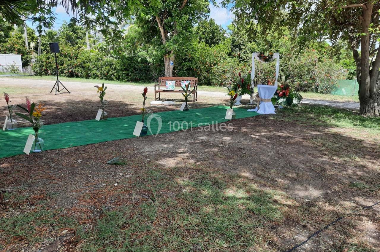 Allée décorée pour une cérémonie en extérieur, bordée de fleurs tropicales, avec une arche et une table en arrière-plan.