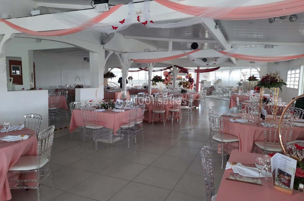 Salle de réception décorée avec des nappes roses, des chaises transparentes et des guirlandes suspendues.