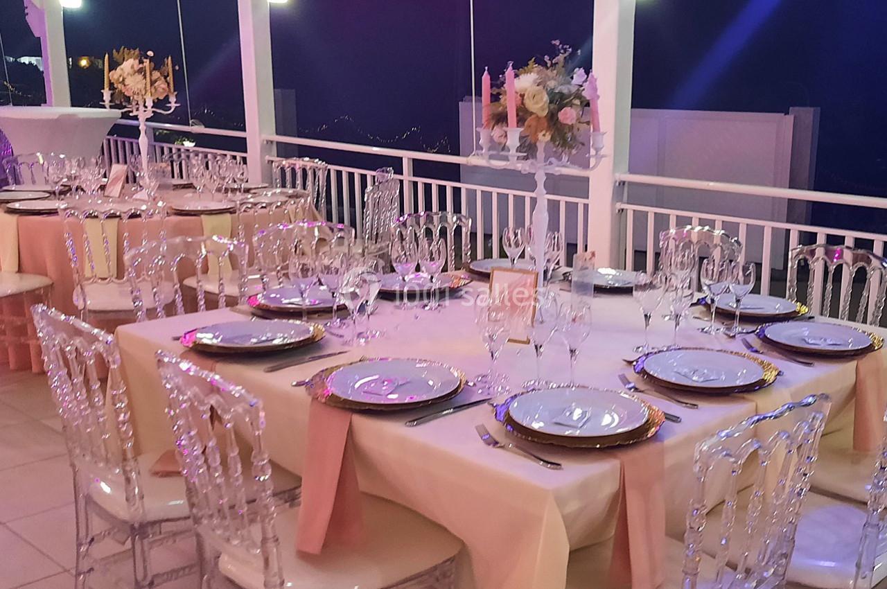 Tables élégamment dressées avec nappes beige, chaises transparentes et décorations florales dans une salle de réception.