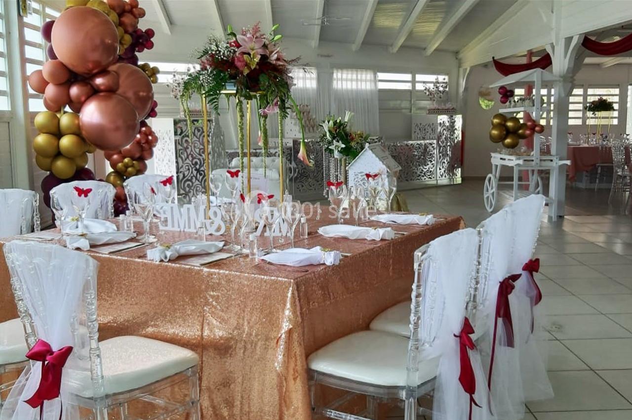 Salle décorée pour une réception avec table recouverte d'une nappe dorée, chaises ornées de nœuds rouges et ballons colorés.