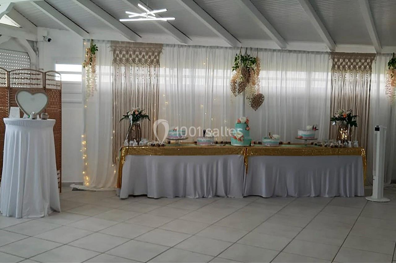 Table décorée pour une réception avec nappes blanches, gâteaux, fleurs et guirlandes lumineuses dans une salle lumineuse.