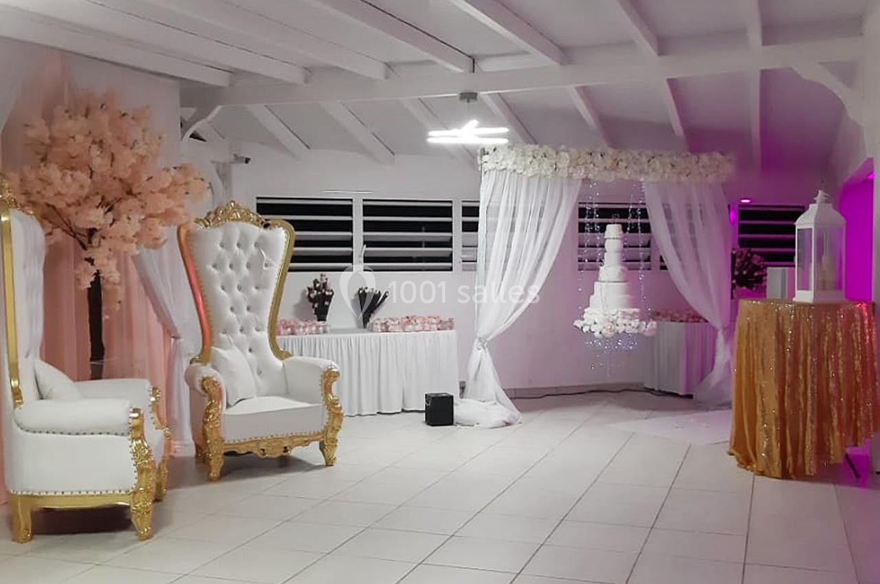 Salle décorée pour un événement avec fauteuils blancs dorés, arche fleurie, table dorée et arbre artificiel rose.