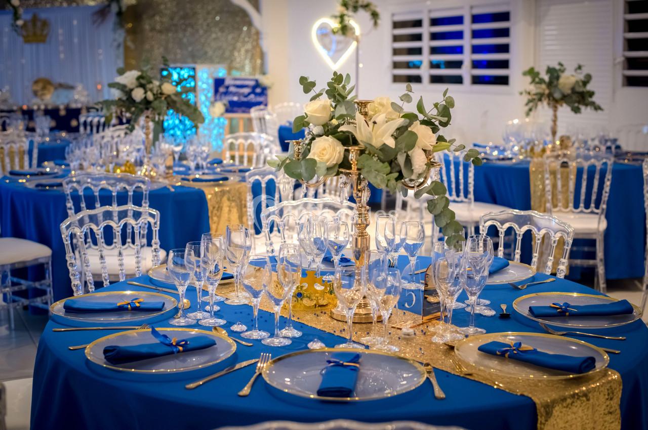 Salle de réception décorée avec des tables rondes ornées de nappes bleues, centres de table floraux et vaisselle élégante.