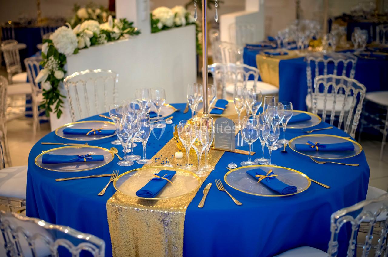 Table élégamment dressée avec nappes bleues, chemin de table doré, vaisselle raffinée et décorations florales blanches.