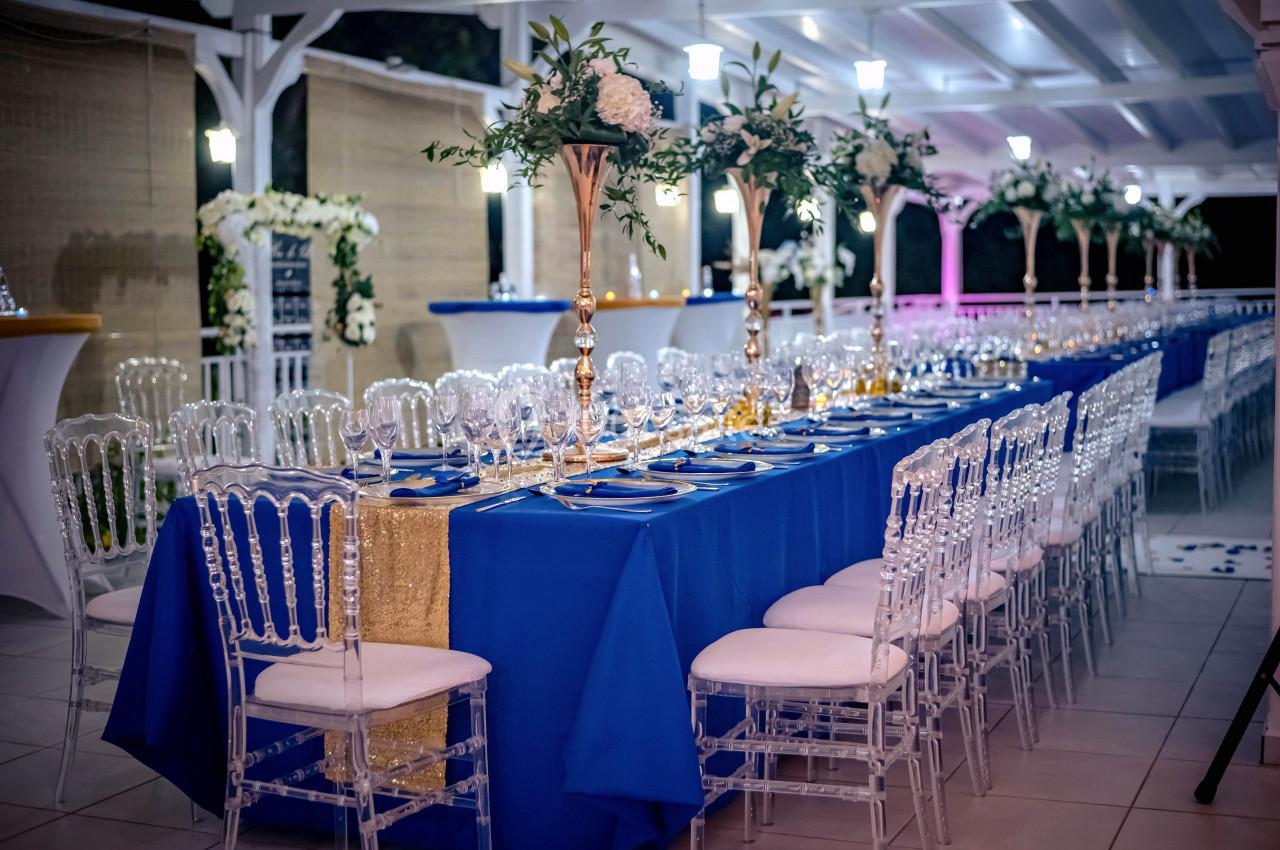 Table de réception élégamment dressée avec nappes bleues, chaises transparentes et décorations florales blanches et dorées.