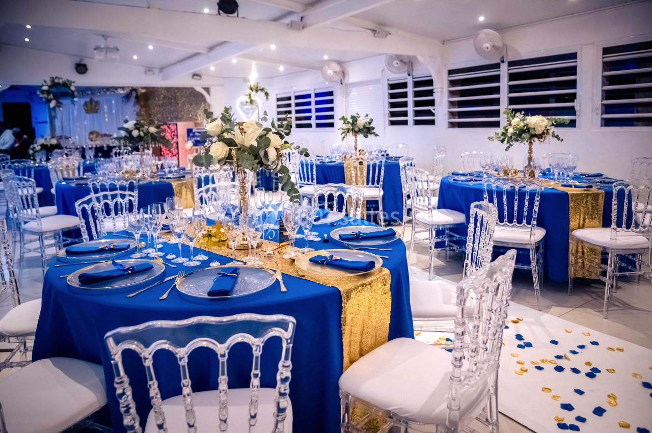 Salle de réception décorée avec des tables dressées en bleu et or, chaises transparentes et arrangements floraux.