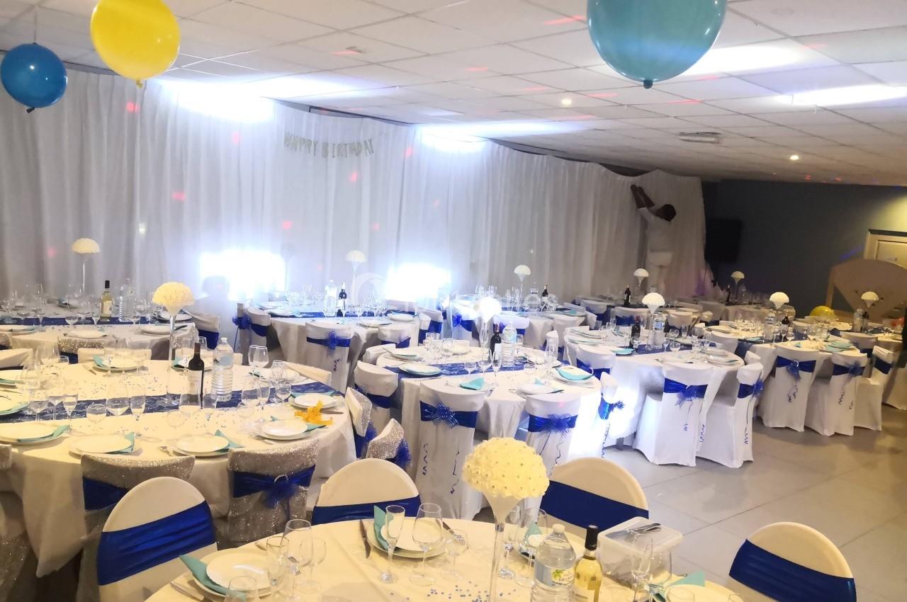 Location salle Bonneuil-sur-Marne (Val-de-Marne) - Salle des Petits Carreaux #1 Salle de réception décorée avec des tables rondes, nappes blanches, rubans bleus et ballons suspendus au plafond.
