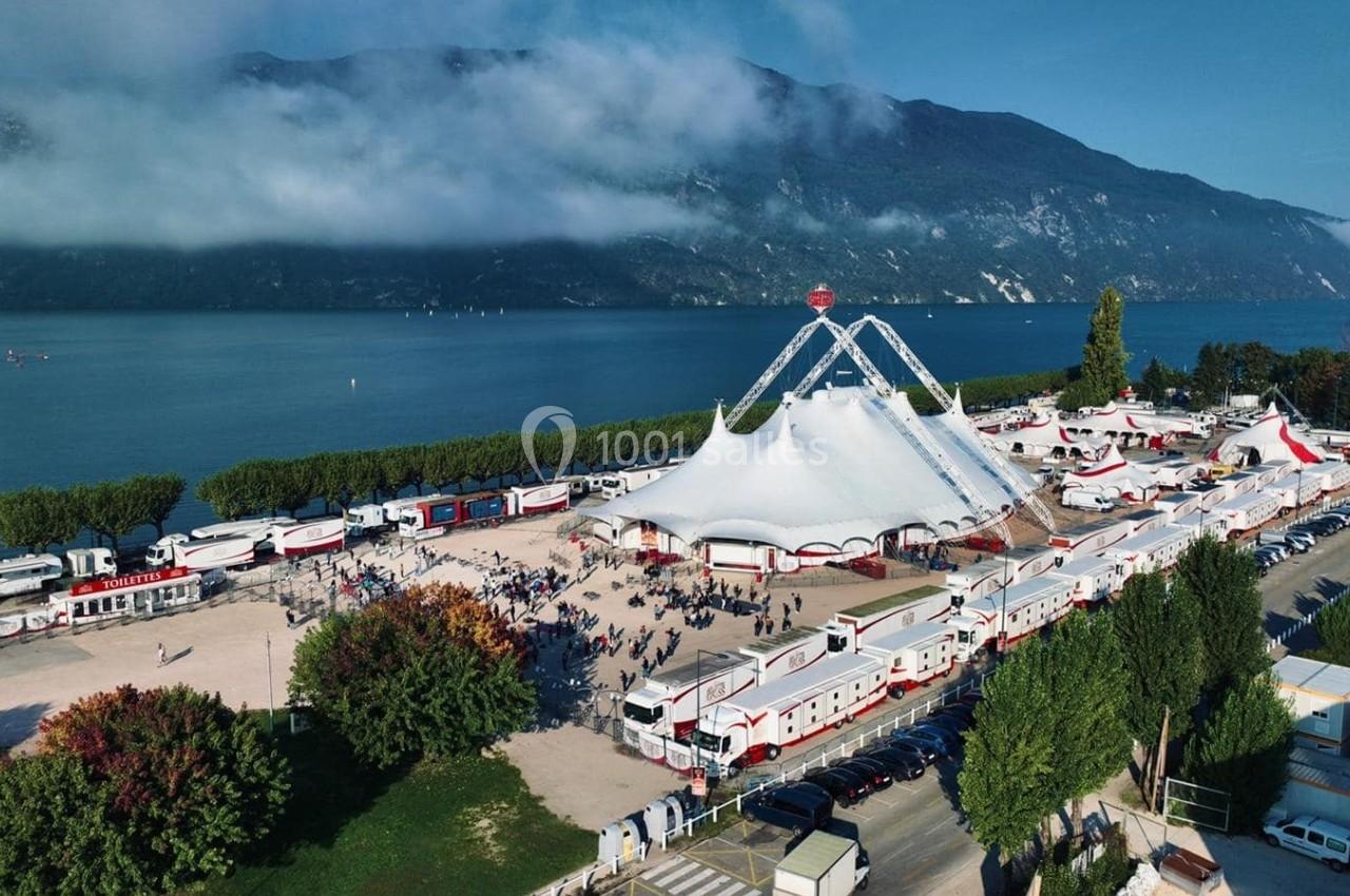Vue aérienne d'un grand chapiteau blanc entouré de caravanes et de visiteurs, près d'un lac et de montagnes.