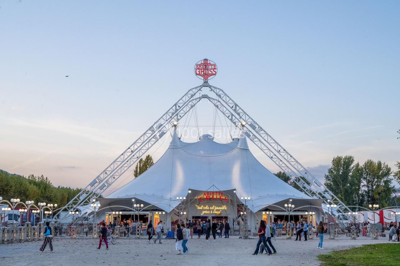 Grand chapiteau blanc d'un cirque avec une structure métallique, entouré de visiteurs en soirée.