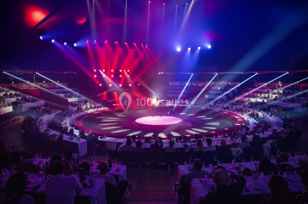 Un artiste sur une scène circulaire éclairée par des projecteurs, entouré de tables avec des spectateurs assis.