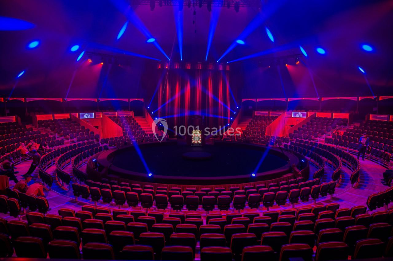 Salle de spectacle circulaire avec sièges rouges, scène centrale éclairée par des projecteurs bleus et rouges.