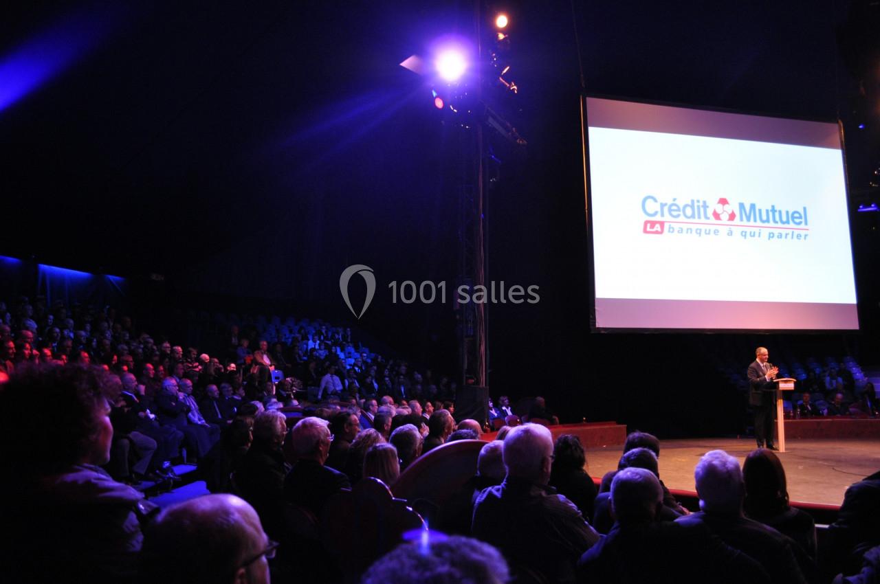 Un orateur s'adresse à un public nombreux dans une salle sombre, avec un écran affichant le logo du Crédit Mutuel.