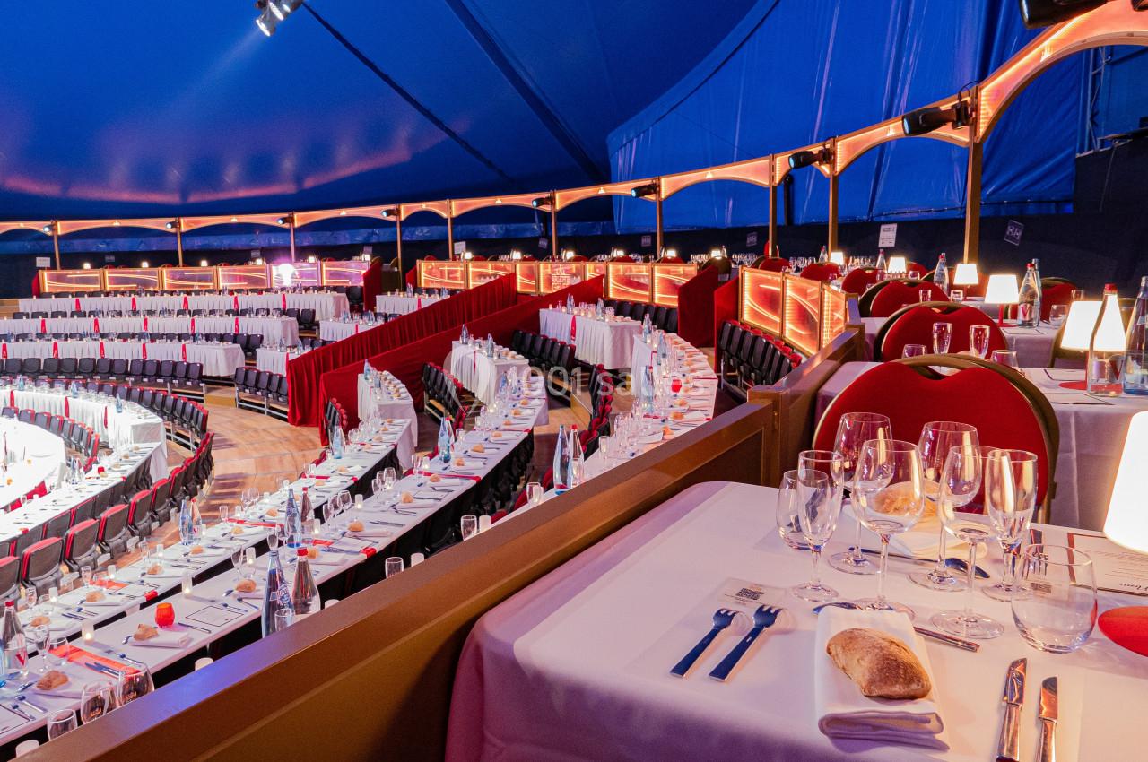 Salle de spectacle sous chapiteau avec tables dressées, nappes blanches et éclairage chaleureux.