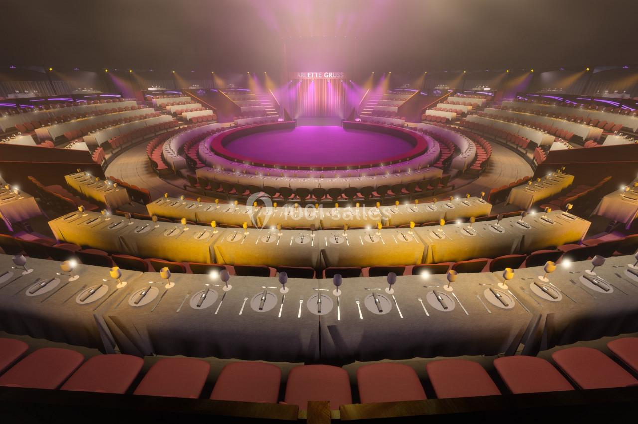 Salle circulaire avec scène centrale, tables dressées en premier plan et gradins illuminés en arrière-plan.
