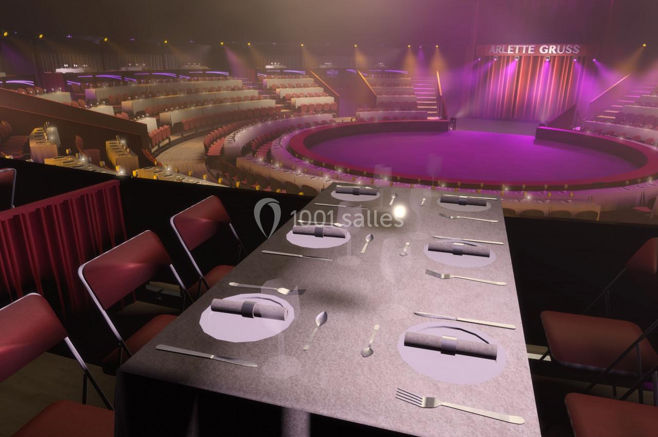 Salle de spectacle circulaire avec scène éclairée, tables dressées au premier plan et gradins en arrière-plan.