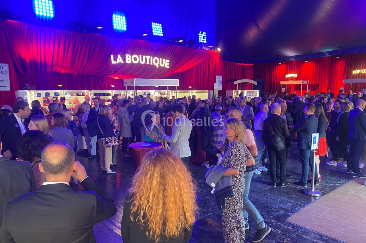 Foule rassemblée dans un espace intérieur éclairé, devant une boutique avec des rideaux rouges en arrière-plan.